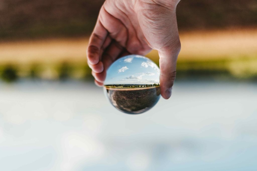 Foto von einer Hand, die über dem Meer eine Glaskugel hält, durch die eine Landschaft und den Himmel gesehen werden können